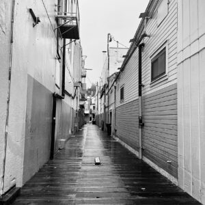 black & white photo of a narrow alley in a city that is laden with wood planks in place of gravel or cement.